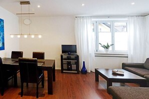A living room with a dining area, a television on a stand, a blue vase, and a window with curtains. - At the spa park - Apartment 1 - 60 sqm (Garmisch-Partenkirchen)