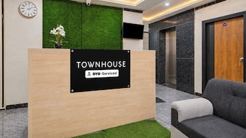 Un mostrador de recepción con un letrero que indica servicio OYO, zona de recepción con un reloj, televisor de pared y un elemento decorativo de césped artificial verde.