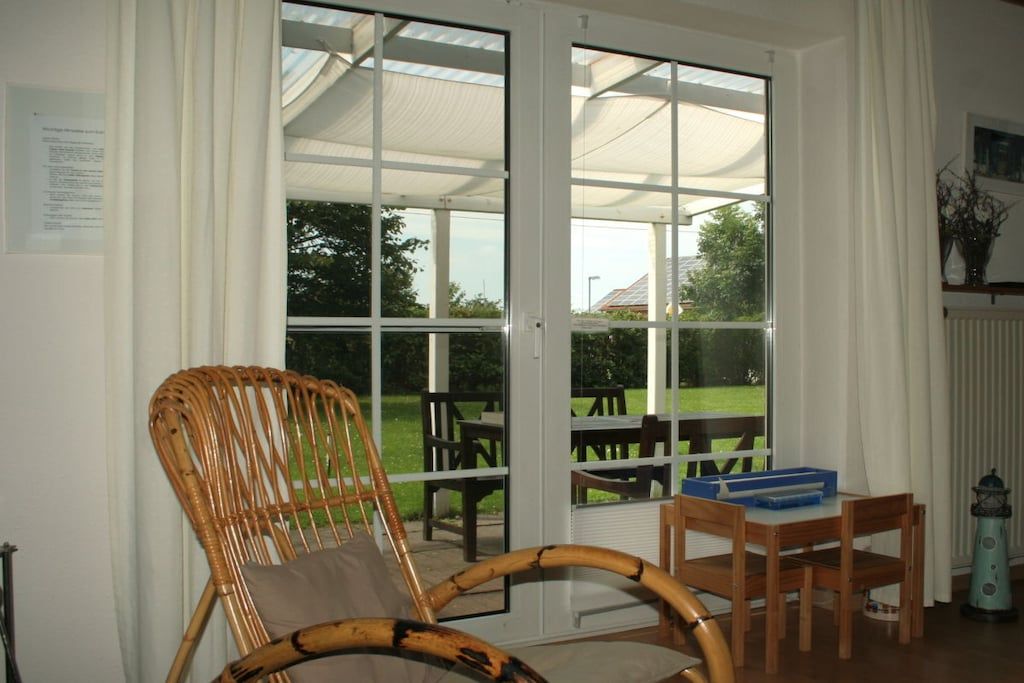 Une pièce avec un fauteuil en osier, une table en bois et une vue, à travers des portes vitrées, sur un espace extérieur aménagé avec des tables et des chaises.