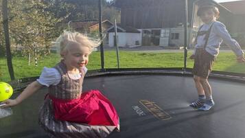 Zwei Kinder spielen auf einem Trampolin mit Netzschutz, umgeben von einer Rasenfläche, mit einem Gebäude im Hintergrund.