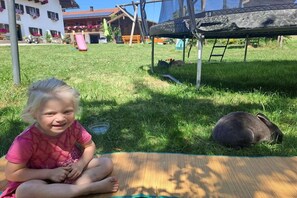 A child sitting on a mat in a grassy outdoor area with a trampoline and a slide in the background.