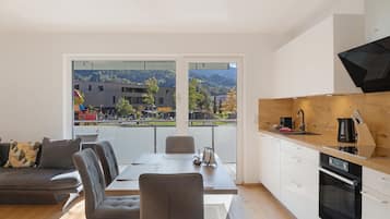 Een moderne woonkamer met een eethoek, grote ramen met uitzicht op een gebouw en heuvels, een keuken met een zwarte kookplaat, en een grijze bank.