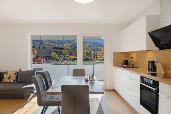 A modern living room with a dining area, large windows with a view of a building and hills, a kitchen with a black stove, and a gray sofa.