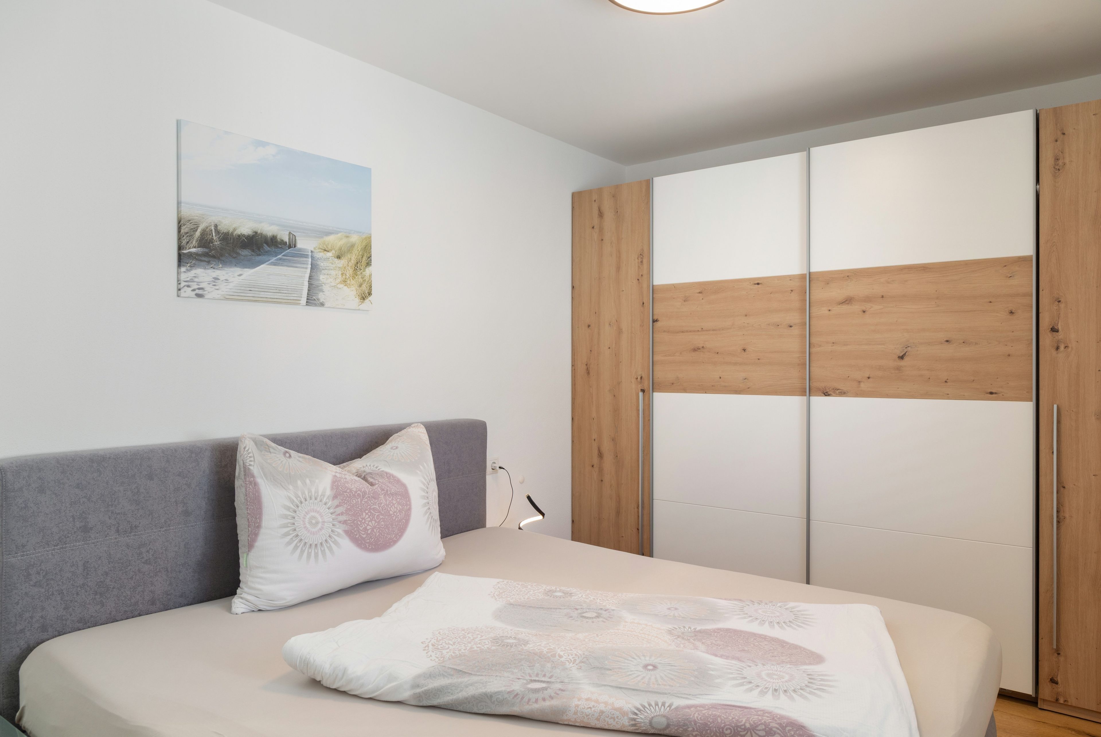 Een slaapkamer met een bed, een kussen, een nachtkastje, een lamp, een houten garderobe en een aan de muur hangend schilderij.