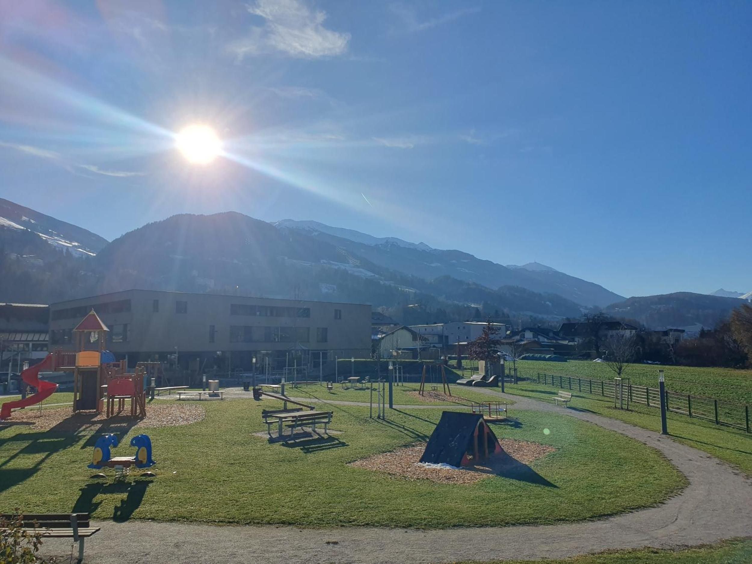 Ein Spielplatz mit verschiedenen Spielgeräten, ein Picknickbereich und ein Weg zu einem Gebäude mit Blick auf das Gebirge.