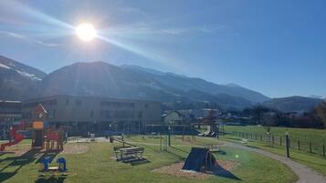 A playground with various play structures, a picnic area, and a path leading to a building with a mountain backdrop.