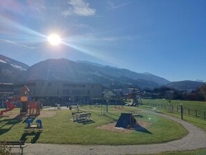 A playground with various play structures, a picnic area, and a path leading to a building with a mountain backdrop.