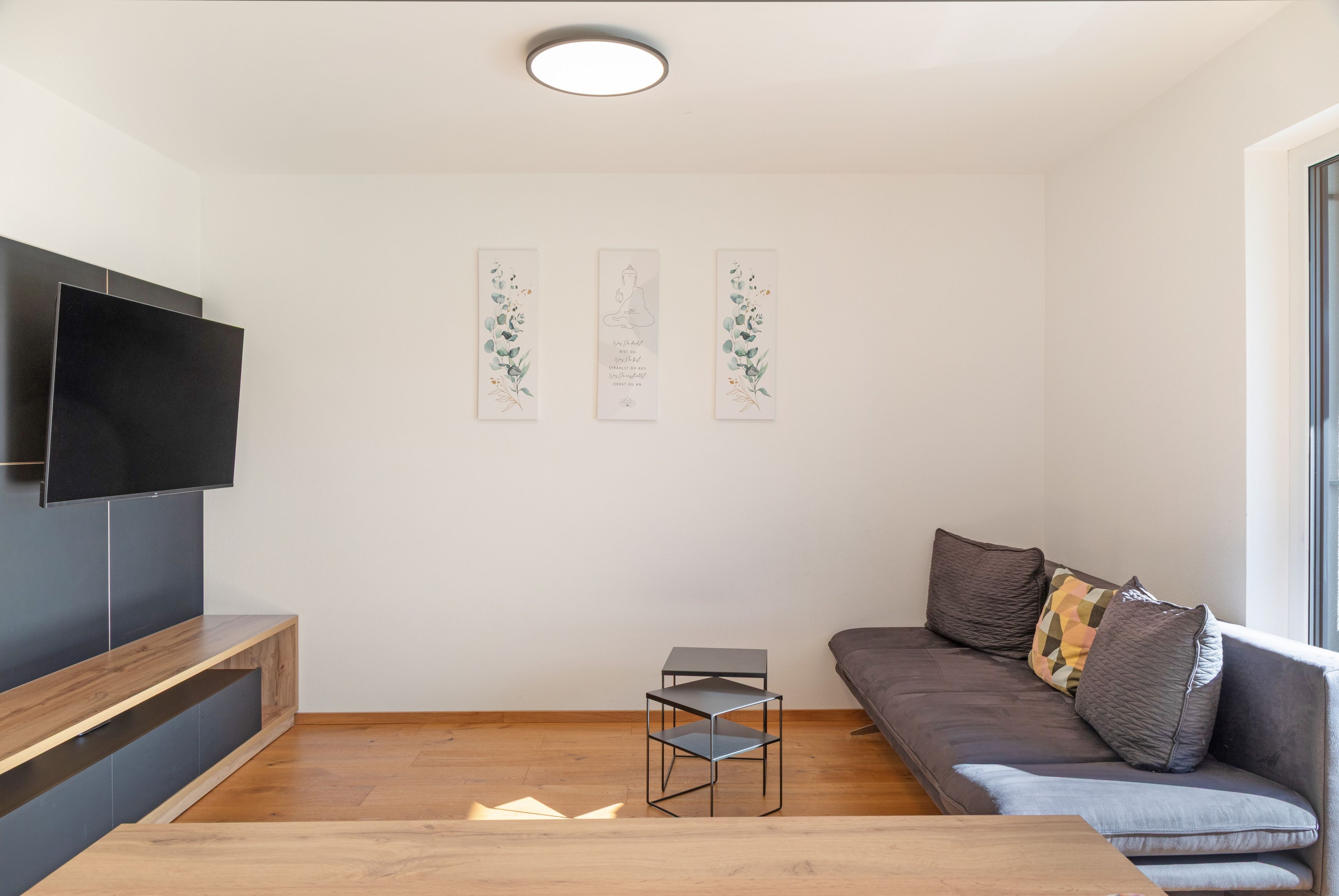 A modern living room with a flat-screen TV, a grey sofa with cushions, a small table, and three wall-mounted artworks.