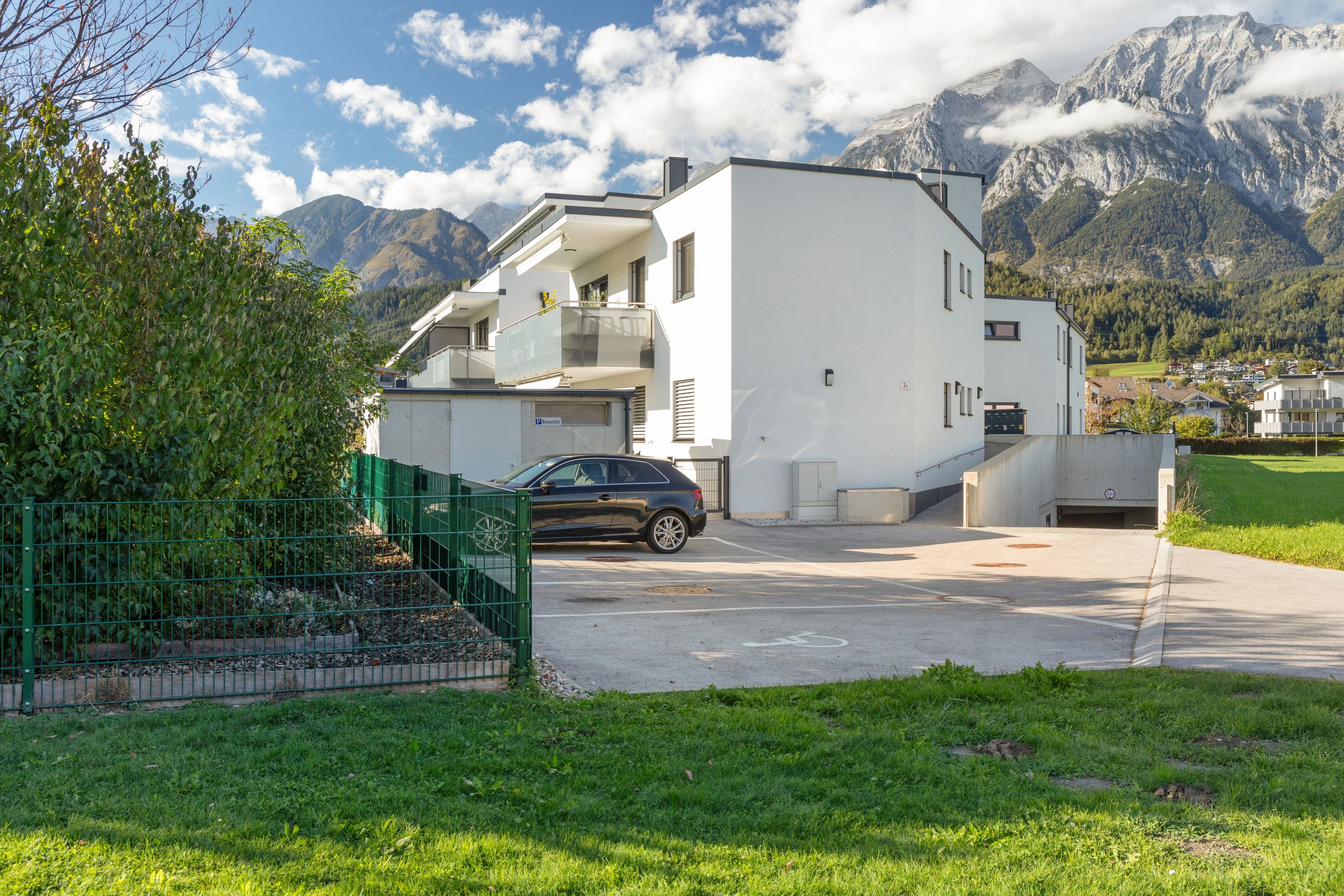 Um edifício moderno de dois andares com um carro estacionado na frente, cercado por vegetação e montanhas ao fundo.