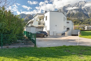 A modern two-story building with a car parked in front, surrounded by greenery and mountains in the background.