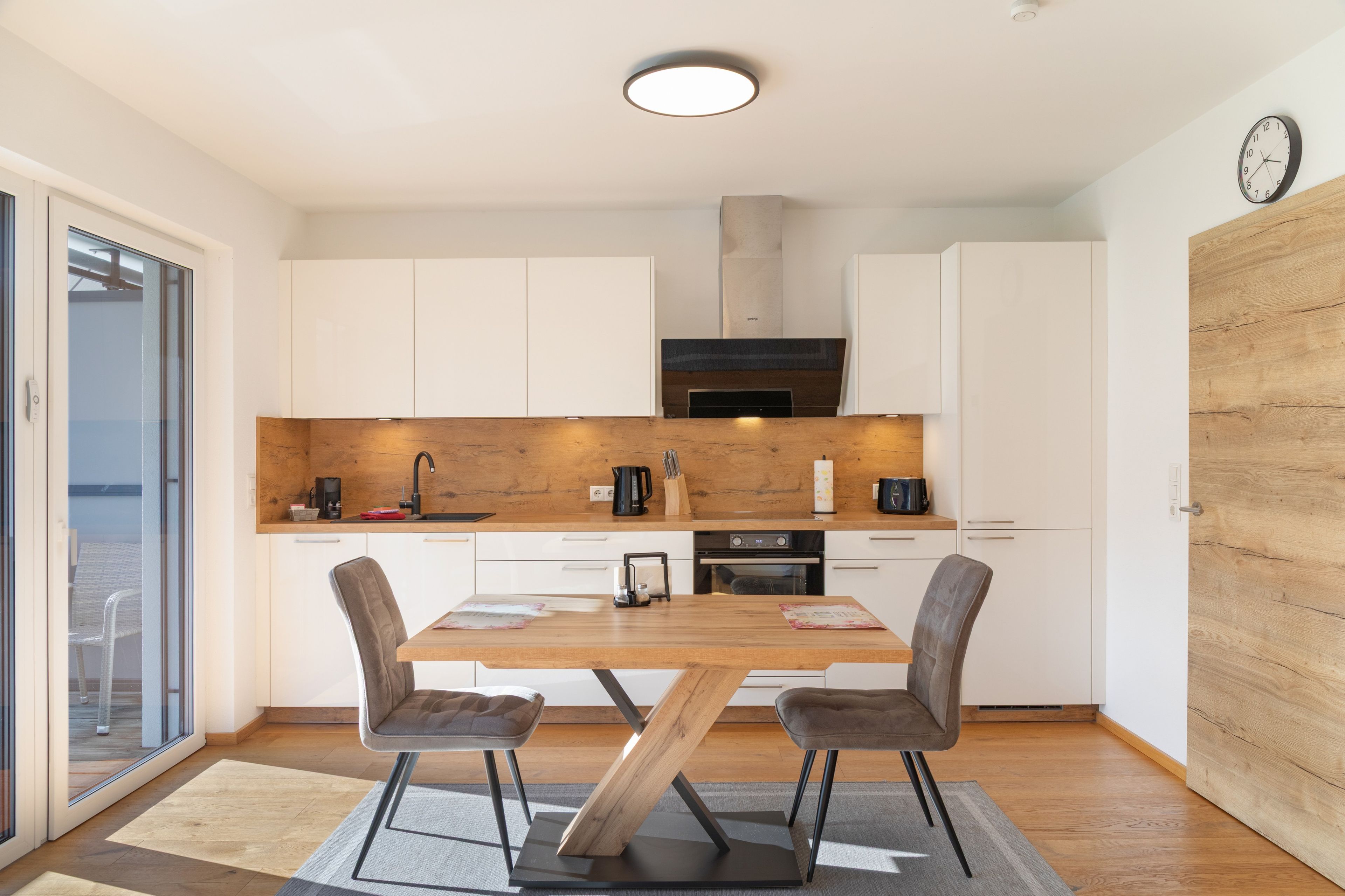 A modern kitchen with white cabinets, a wooden dining table, two chairs, and a built-in oven.