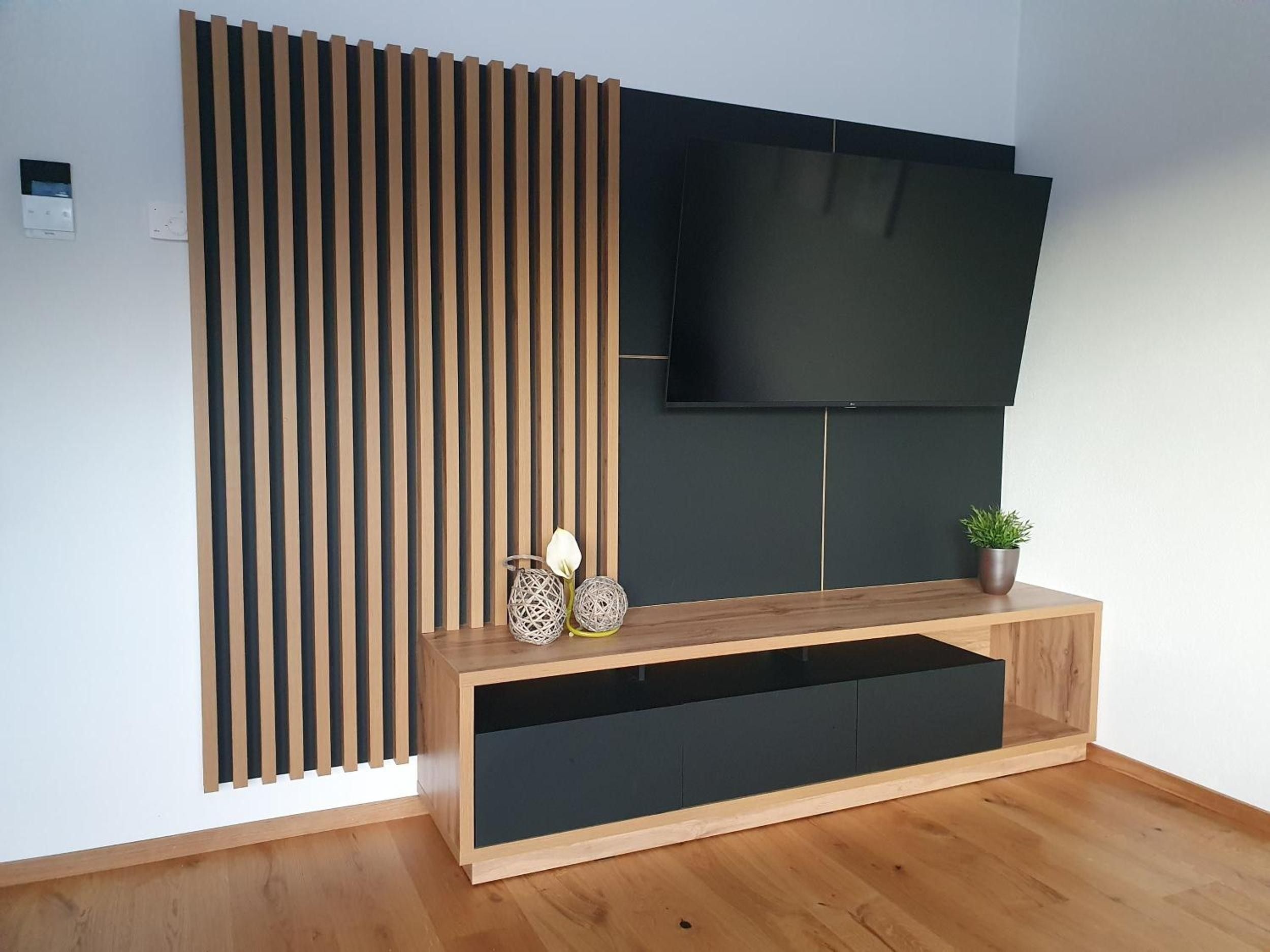 Ein modernes Wohnzimmer mit einem flachbildfernseher, der an einer dunklen Wand angebracht ist, einer Trennwand aus Holzlamellen und einem Holztische mit Dekorationselementen.