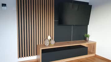 Ein modernes Wohnzimmer mit einem flachbildfernseher, der an einer dunklen Wand angebracht ist, einer Trennwand aus Holzlamellen und einem Holztische mit Dekorationselementen.