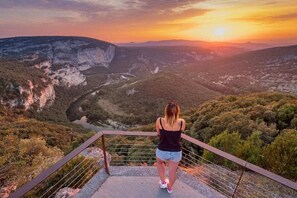 A person standing on a viewing platform overlooking a valley with a river, with a sunset in the background. - Gite Marie -Mas Paraloup (Lagorce)