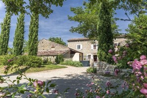 A stone house with a gravel driveway, surrounded by tall trees and flowering plants. - Gite Marie -Mas Paraloup (Lagorce)