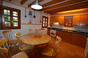 Una cocina con armarios de madera, una mesa redonda de madera y sillas. Se ve un ventanal con vistas a la vegetación.