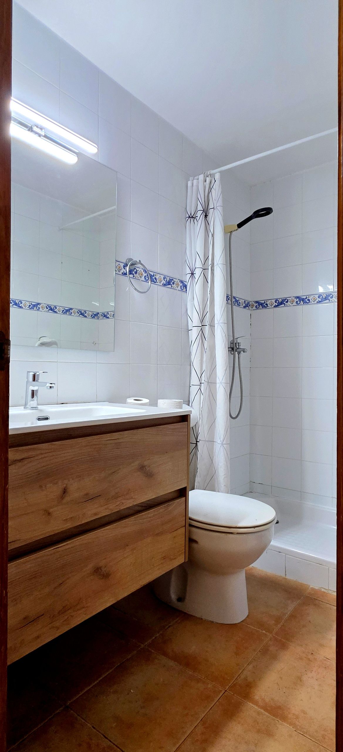 A bathroom with a wooden vanity, a white toilet, a shower with a curtain, and a mirror with a decorative tile border.
