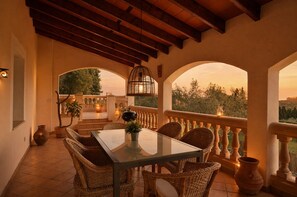 Eine überdachte Terrasse mit Tisch und Korbstühlen, einem hängenden Laternen und Blick auf die Landschaft.