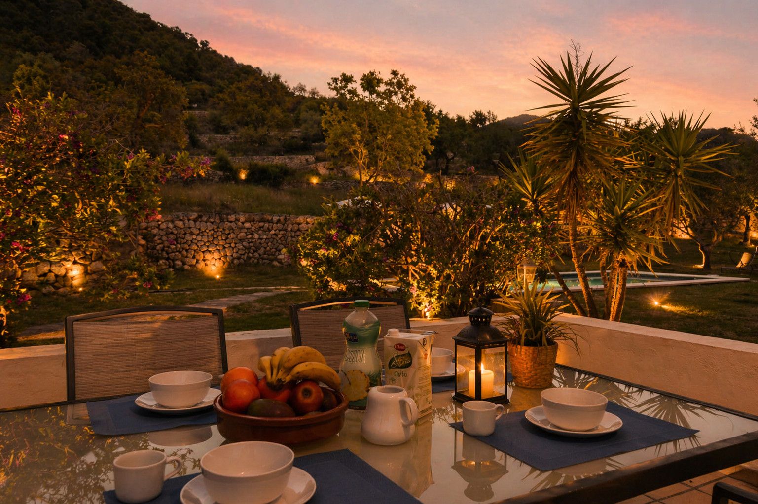 Une table à manger agrémentée d’un panier de fruits, de tasses et d’une bougie, le tout mis en valeur par une vue pittoresque sur une piscine et des arbres au crépuscule.
