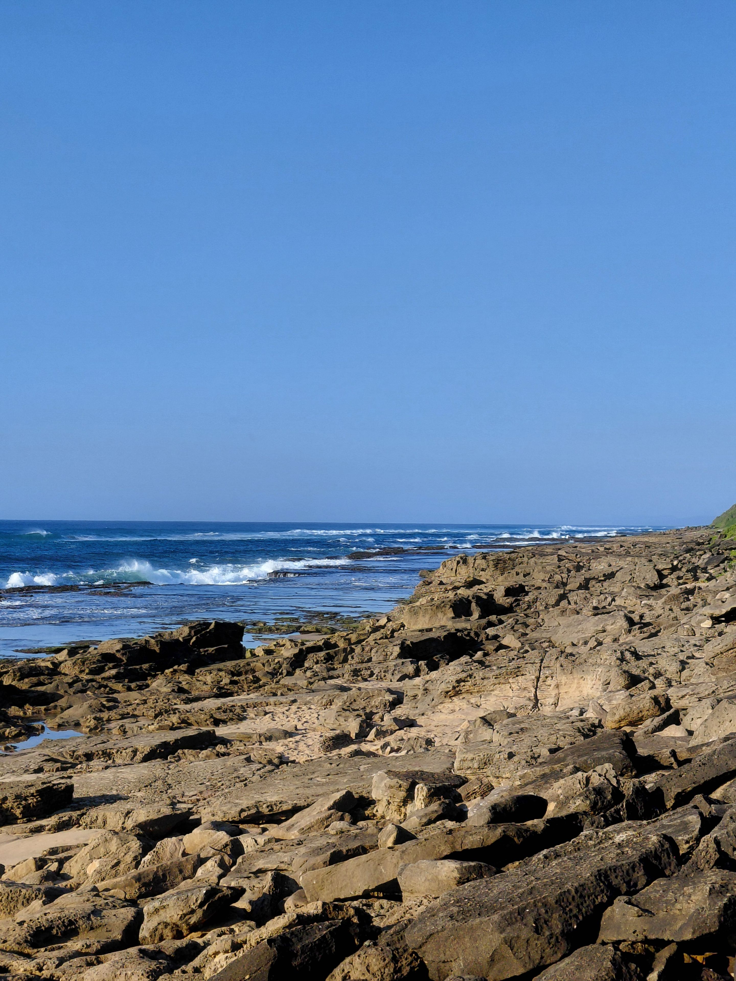 A rocky shoreline with waves crashing against the rocks under a clear blue sky.