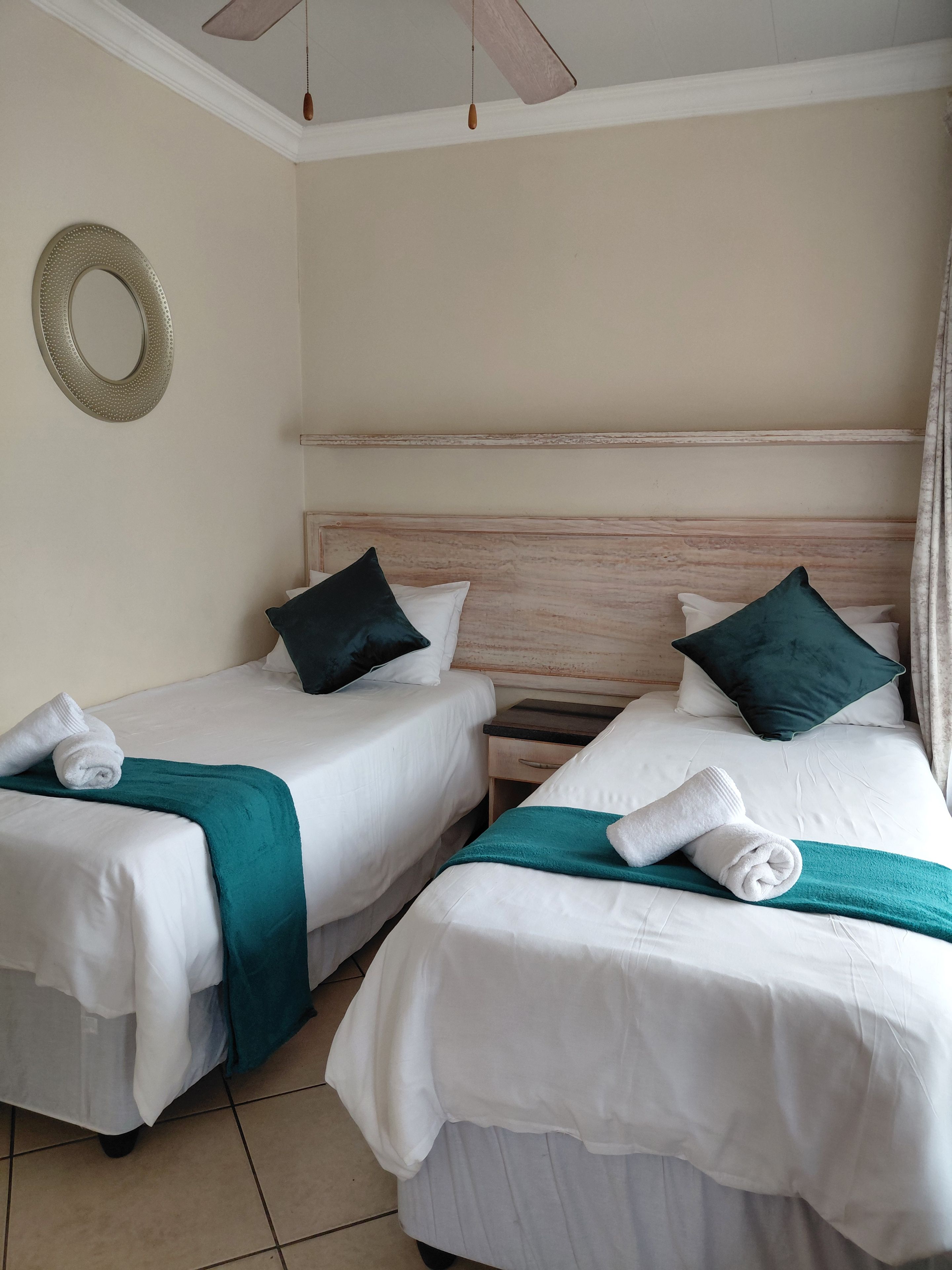 A hotel room with two single beds, each with a teal throw blanket and a rolled white towel. There is a wooden headboard, a nightstand with a drawer, and a wall-mounted mirror.