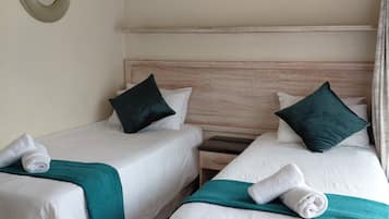 A hotel room with two single beds, each with a teal throw blanket and a rolled white towel. There is a wooden headboard, a nightstand with a drawer, and a wall-mounted mirror.