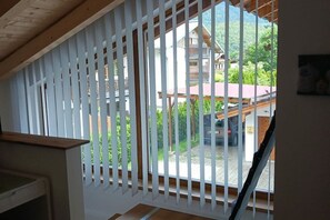 A room with a view through a window with white blinds, a wooden ceiling, and a visible outdoor area with a building and parked cars. - Holiday apartment with 1 bedroom (Schönau am Königssee)