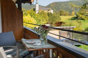 A balcony with wicker furniture, a table with a vase of flowers, and a scenic view of a church and greenery. - Holiday apartment with 2 bedrooms (Ruhpolding)