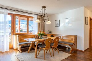 A dining area with a wooden bench, a table, and two chairs. There is a window with curtains, a radiator, and a vase with flowers on the table. - Holiday apartment with 2 bedrooms (Ruhpolding)