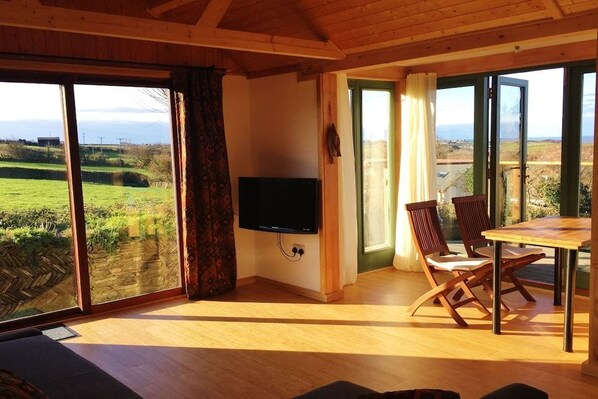 A room with a television, a dining table with chairs, and large sliding glass doors leading to a green landscape.