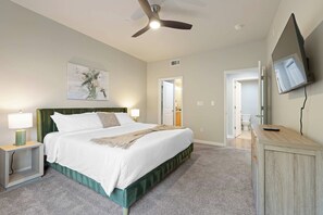 A hotel room with a bed, bedside lamps, a ceiling fan, a painting, a television, and a door leading to a bathroom.