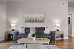Ein Wohnzimmer mit einem dunkelbraunen Ledersofa, zwei Beistelltischen mit Lampen, einem Couchtisch und einem Gemälde an der Wand.