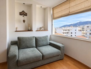 A grey sofa in a room with a large window, wooden floor, and decorative items on shelves. - Nini's Maison - Terrace Living Above Kalamata City (Kalamata)