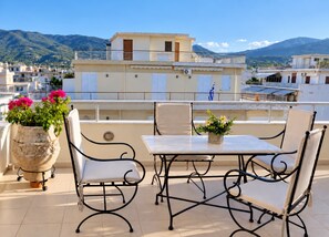 A terrace with a table and chairs, potted plants, and a view of buildings and mountains. - Nini's Maison - Terrace Living Above Kalamata City (Kalamata)