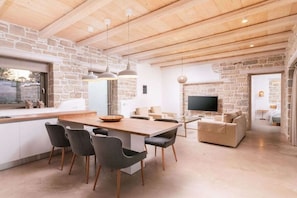 Ein modernes Interieur mit einer Steinwand, einer Holzbalkendecke, einem Essbereich mit Stühlen und Tisch, einer Couch und einem Fernseher.