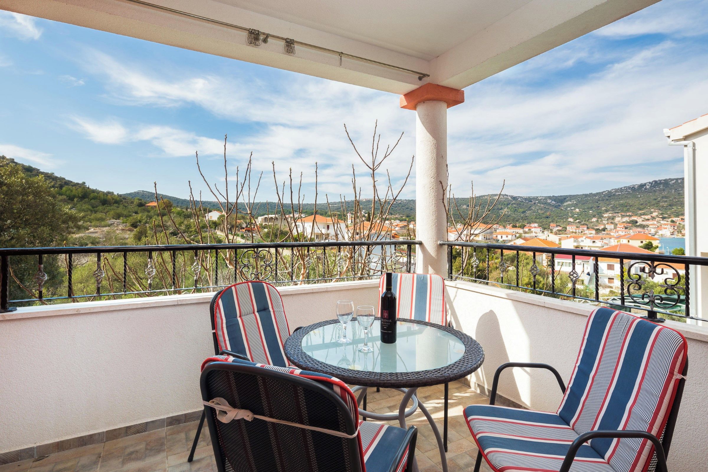 A balcony with a glass table, striped chairs, and a bottle of wine.
