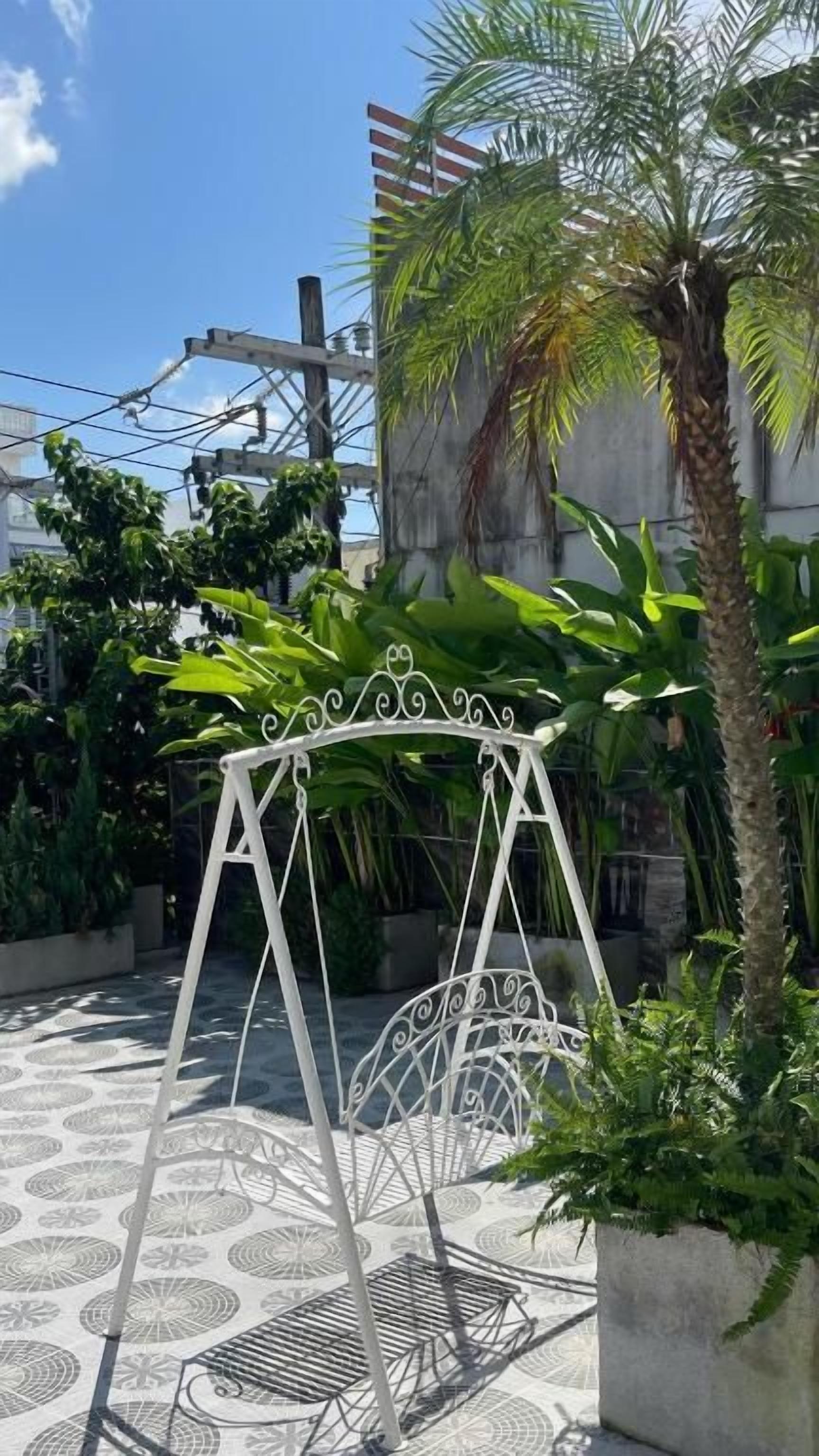 Un columpio metálico en un patio con piso estampado, rodeado de follaje y una palmera.