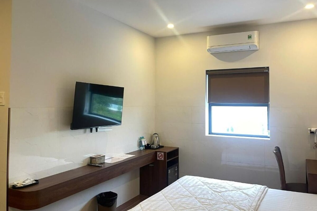 A hotel room with a bed, a TV mounted on the wall, a window with a view, a desk with a trash bin, and an air conditioning unit.