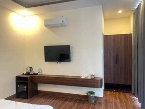 Habitación de hotel con cama, televisor de pantalla plana montado en la pared, banco de madera, mesita con tetera y tazas, y un cubo de basura.