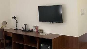 Una stanza con una TV a schermo piatto fissata al muro, una scrivania in legno con una lampada e un piccolo frigorifero.