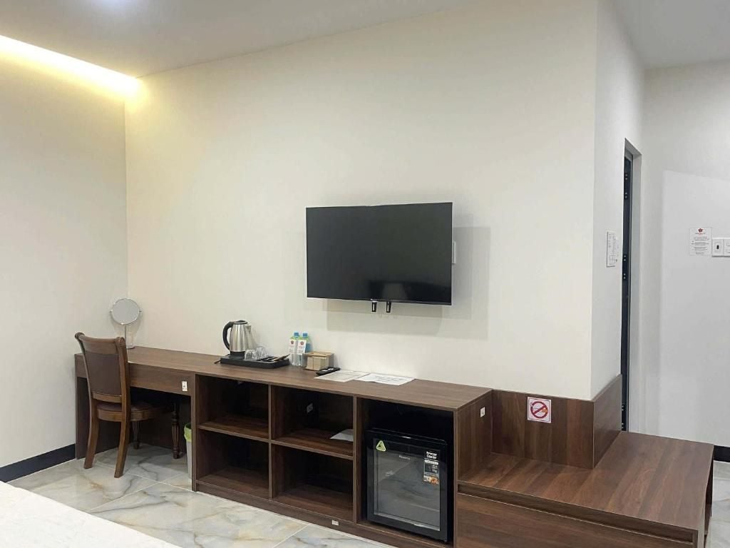 Una habitación con televisión de pantalla plana montada en la pared, un escritorio de madera con silla, un refrigerador pequeño y una cafetera.