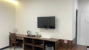 Una habitación con televisión de pantalla plana montada en la pared, un escritorio de madera con silla, un refrigerador pequeño y una cafetera.