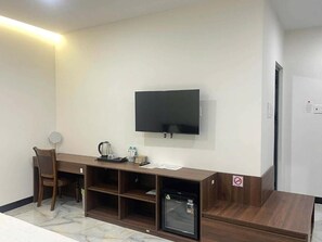 Una habitación con televisión de pantalla plana montada en la pared, un escritorio de madera con silla, un refrigerador pequeño y una cafetera.