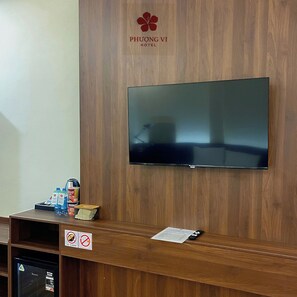 Zona de recepción de un hotel con un televisor de pantalla plana montado en una pared de madera, un refrigerador y una pequeña mesa con agua embotellada y una cafetera.