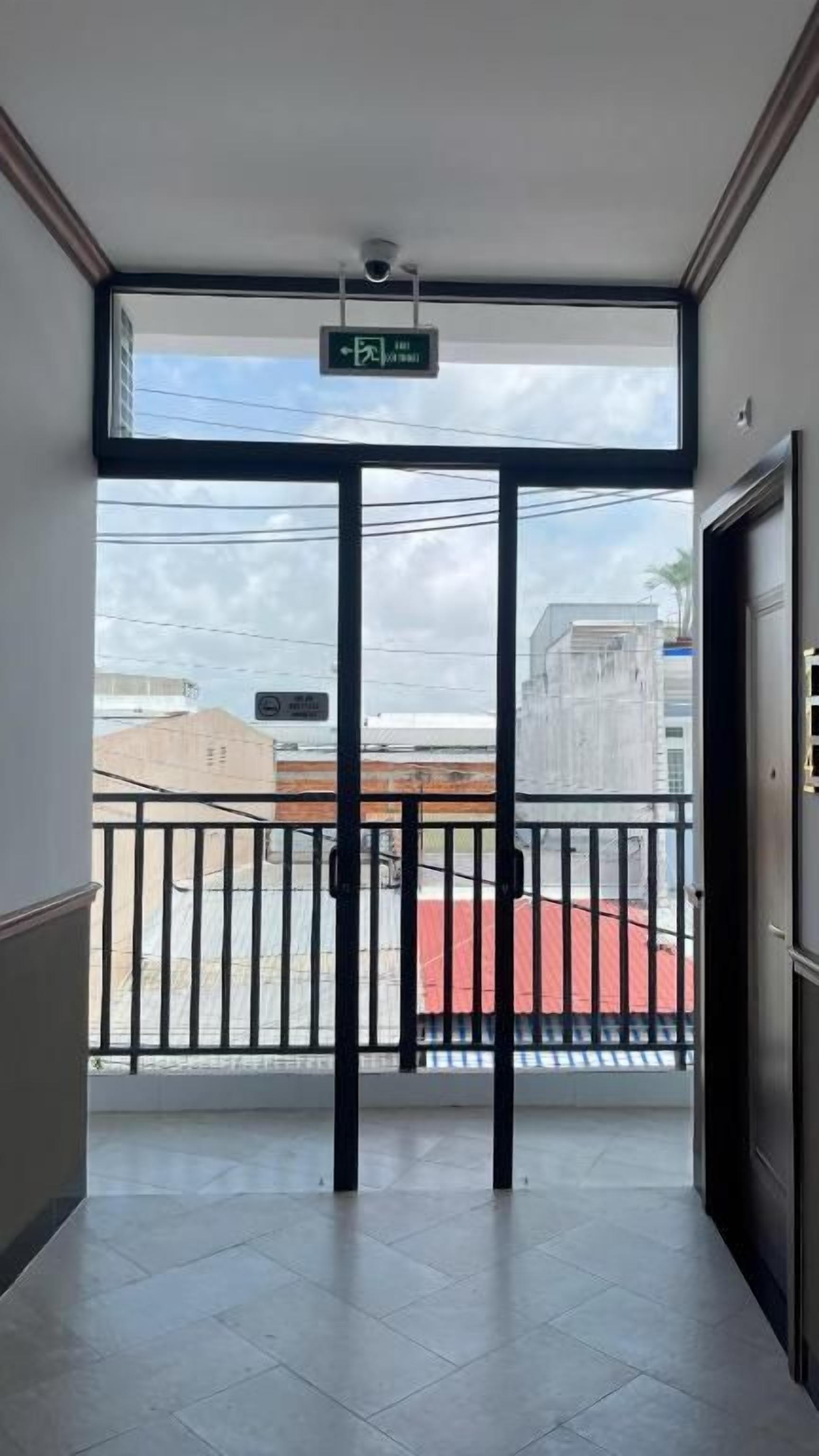 A hotel entrance with a glass door, a balcony with a red mat, and a view of buildings and a clear sky.