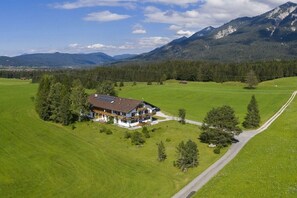 Ein Haus mit geneigtem Dach, umgeben von einem grünen Feld und einem Wald, mit Bergen im Hintergrund.