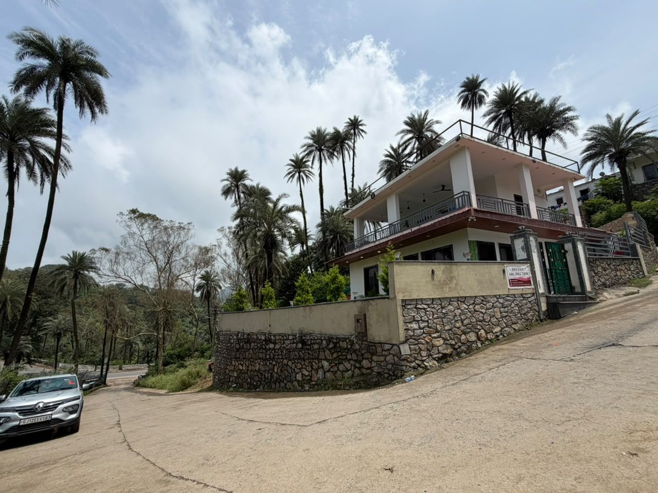 A two-story house with a balcony, surrounded by palm trees and a stone wall.