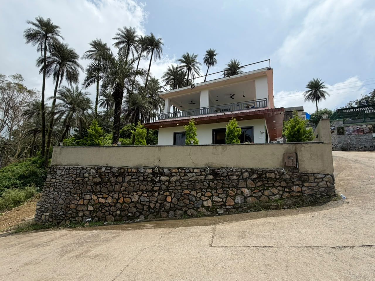 A two-story building with a stone retaining wall, palm trees in the background, and a clear sky.