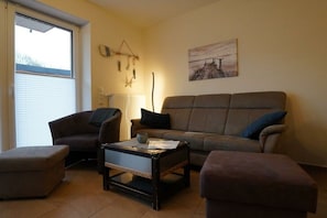 A living room with a brown sofa, a brown armchair, a coffee table, and a window with blinds. - Holiday apartment with 2 bedrooms (Norddeich)
