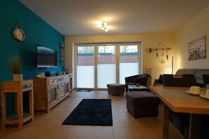 Ein Wohnzimmer mit Fernseher, Sofa, Hocker und Esstisch.